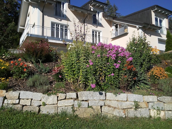 A multi-story building with balconies, surrounded by a stone wall and various flowering plants.