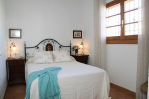 A neatly made bed with a white bedspread and two pillows, flanked by two bedside tables with lamps and framed pictures. A window with wooden shutters is visible in the background. - Lovely apartment for 4 guests with A/C, WIFI, TV and terrace (Chipiona)
