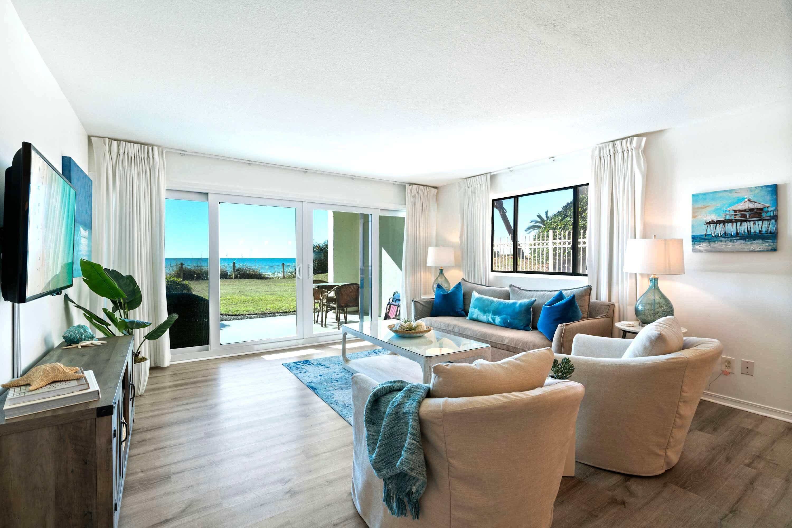 A spacious living room with a large window overlooking a beach, a flat-screen TV, and a painting of a pier.