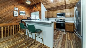 Una cocina con pisos de madera, una isla central con taburetes de bar, gabinetes de madera y electrodomésticos de acero inoxidable, y un techo inclinado.