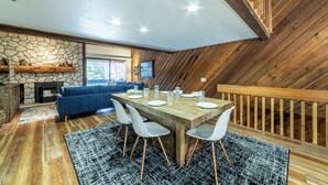 Un comedor con mesa y sillas de madera, chimenea de piedra y un sofá azul en una habitación con paredes de madera y techo inclinado.