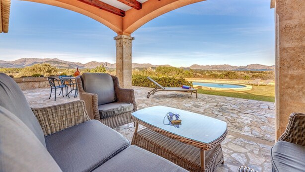 Un patio techado con mobiliario de mimbre, una mesa de piedra y vistas a una piscina y montañas.