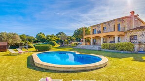 Una piscina rodeada por un césped cuidado, un edificio de piedra con balcones, y un cielo azul despejado.