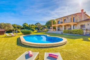 A swimming pool surrounded by a well-manicured lawn, a stone building with balconies, and a clear blue sky.