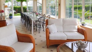 A sunlit dining area with a long table and chairs, surrounded by large windows offering a view of the garden. - Ca Romeu by Interhome (Cambrils)