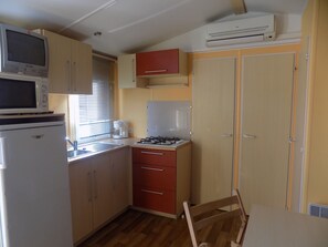 A compact kitchen with a microwave, stove, and refrigerator. There is a window with blinds, a sink, and a chair.