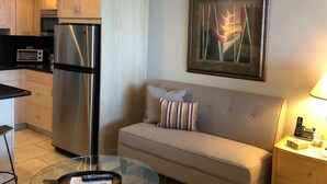 Un espacio habitable pequeño con kitchenette, un sofá con almohadones a rayas, una mesa de centro de vidrio y una obra de arte enmarcada en la pared.