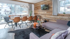 Ein modernes Wohnzimmer mit einer Holzvertäfelung, einem großen Fenster, einer Couch, Sesseln, einem Esstisch und einem Flachbildfernseher.