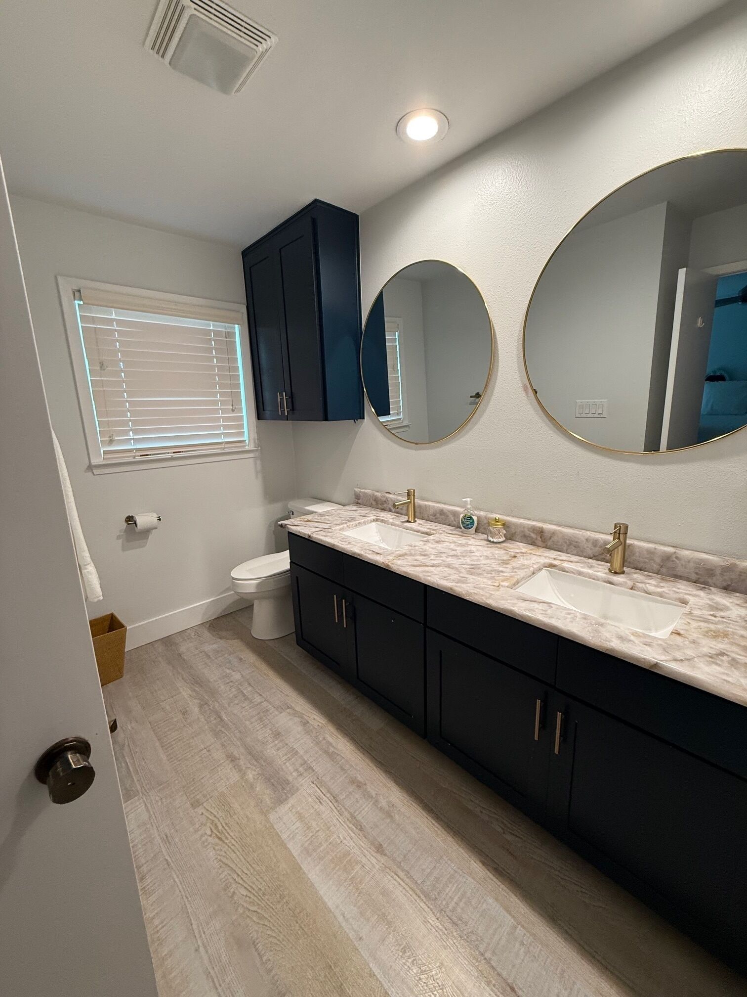 A modern bathroom with two sinks, dark cabinets, a toilet, and a window with blinds.