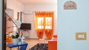 Una habitación con televisión, cortinas naranjas, un sofá blanco, una mesita con una planta y una botella, y un letrero en la pared.