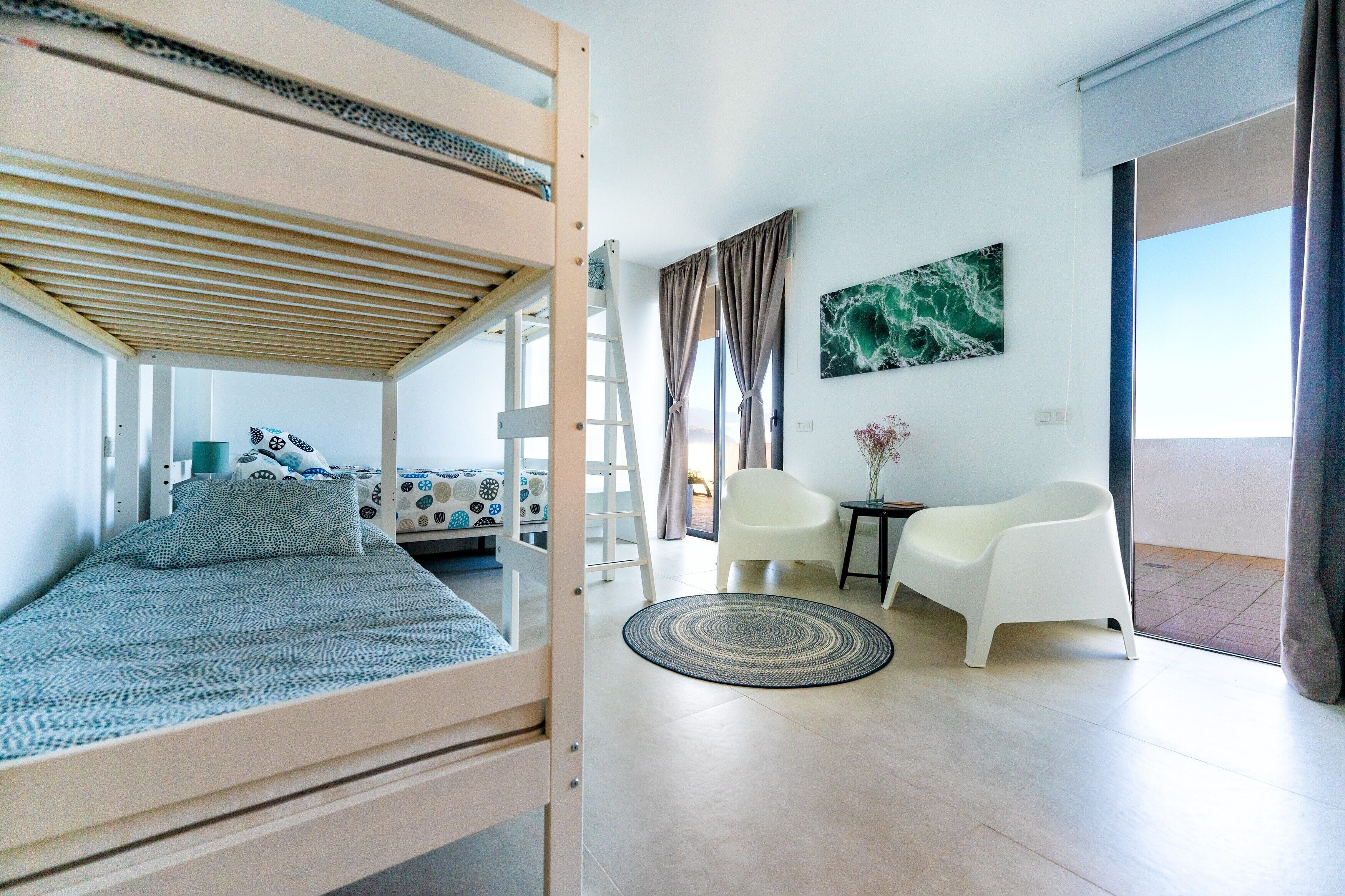 A room with a bunk bed, a white chair, a small table with a vase, a painting on the wall, and a view through a door.