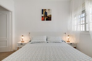 Un dormitorio con una cama, dos mesitas de noche con lámparas, una obra de arte colgada en la pared y un ventanal con cortinas translúcidas.