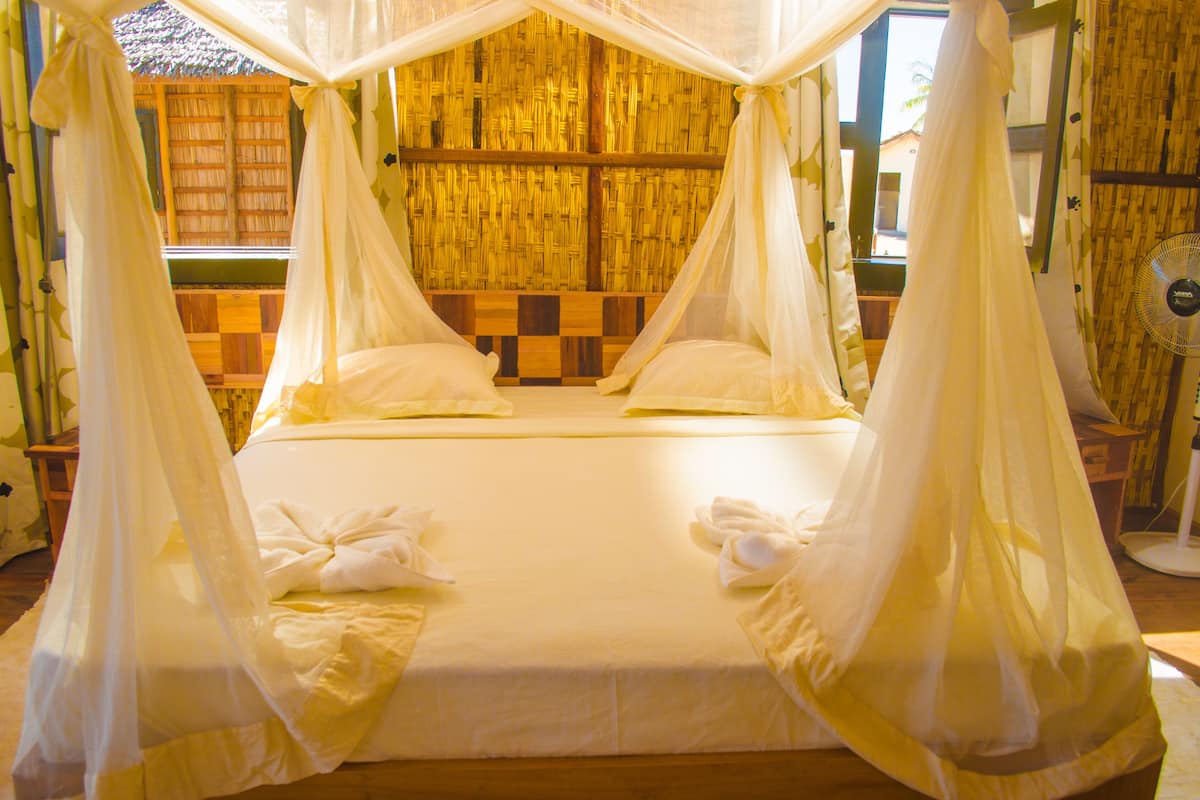 A four-poster bed with white linens and sheer curtains, a wooden headboard, and a view of a bamboo wall.