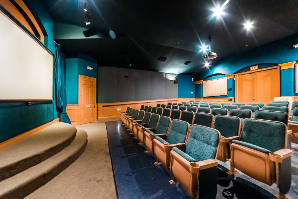 A theater with rows of green upholstered seats, a large screen, and an exit door labeled "EXIT 1".
