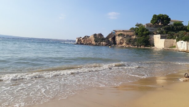 Ein Strand mit klarem Wasser, sandigem Ufer und felsigen Klippen mit Gebäuden darauf.