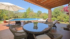 Un patio al aire libre con mobiliario de mimbre, una mesa de vidrio, un parasol grande y una piscina al fondo.