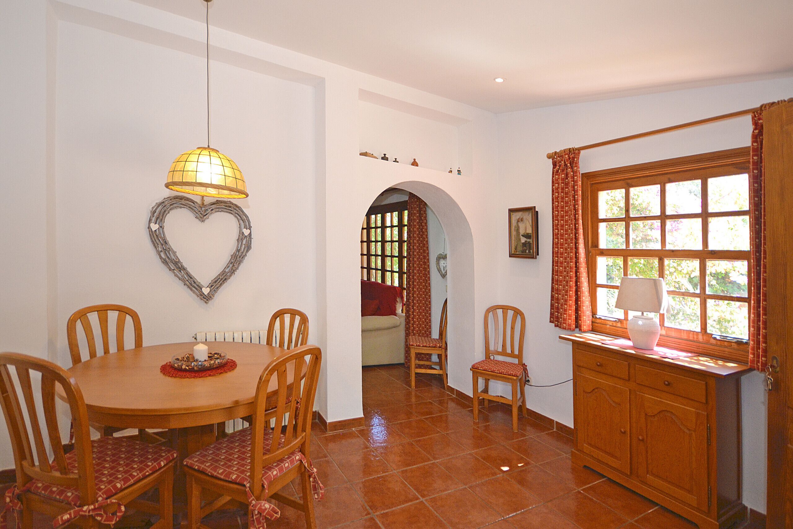 Un comedor con una mesa redonda, sillas, un adorno en forma de corazón en la pared, una lámpara colgante y una ventana con cortinas.