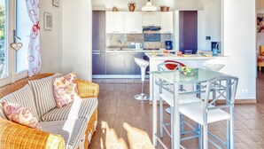 Un salon chaleureux avec un canapé en osier, une table à manger en verre entourée de chaises, et une cuisine en arrière-plan dotée d’appareils modernes et d’un plan de travail carrelé.