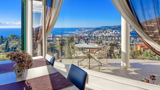 Une terrasse avec une table et des chaises, un vase rempli de fleurs séchées et une vue sur une ville côtière.