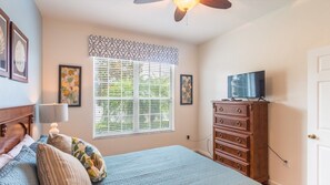Une chambre à coucher comprenant un lit, une commode, un téléviseur et une fenêtre avec des stores.