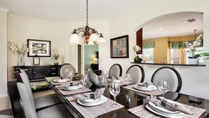 A dining area with a long table set for six, featuring a chandelier, framed artwork, and a view of the kitchen. - New Listing! Windsor Hills Resort, 3mi to Disney & 12mi to Universal - GRH438788 (Kissimmee)