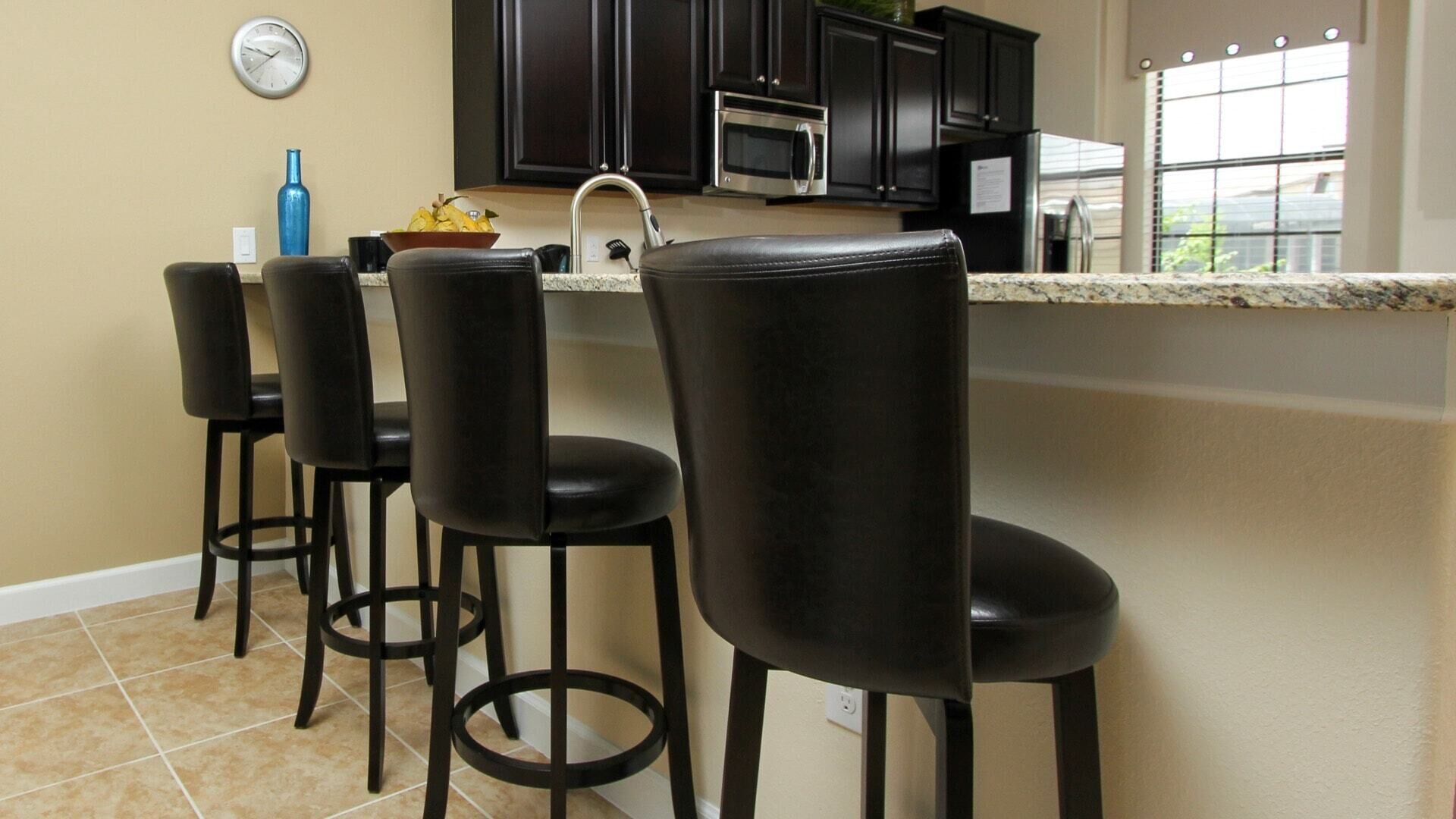 A kitchen with a bar area featuring four black bar stools, a stainless steel microwave, and a window with blinds.
