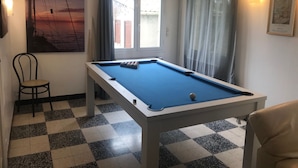 Une table de billard dans une pièce dont le sol est à carreaux, avec une chaise, une fenêtre avec des rideaux et un tableau accroché au mur.
