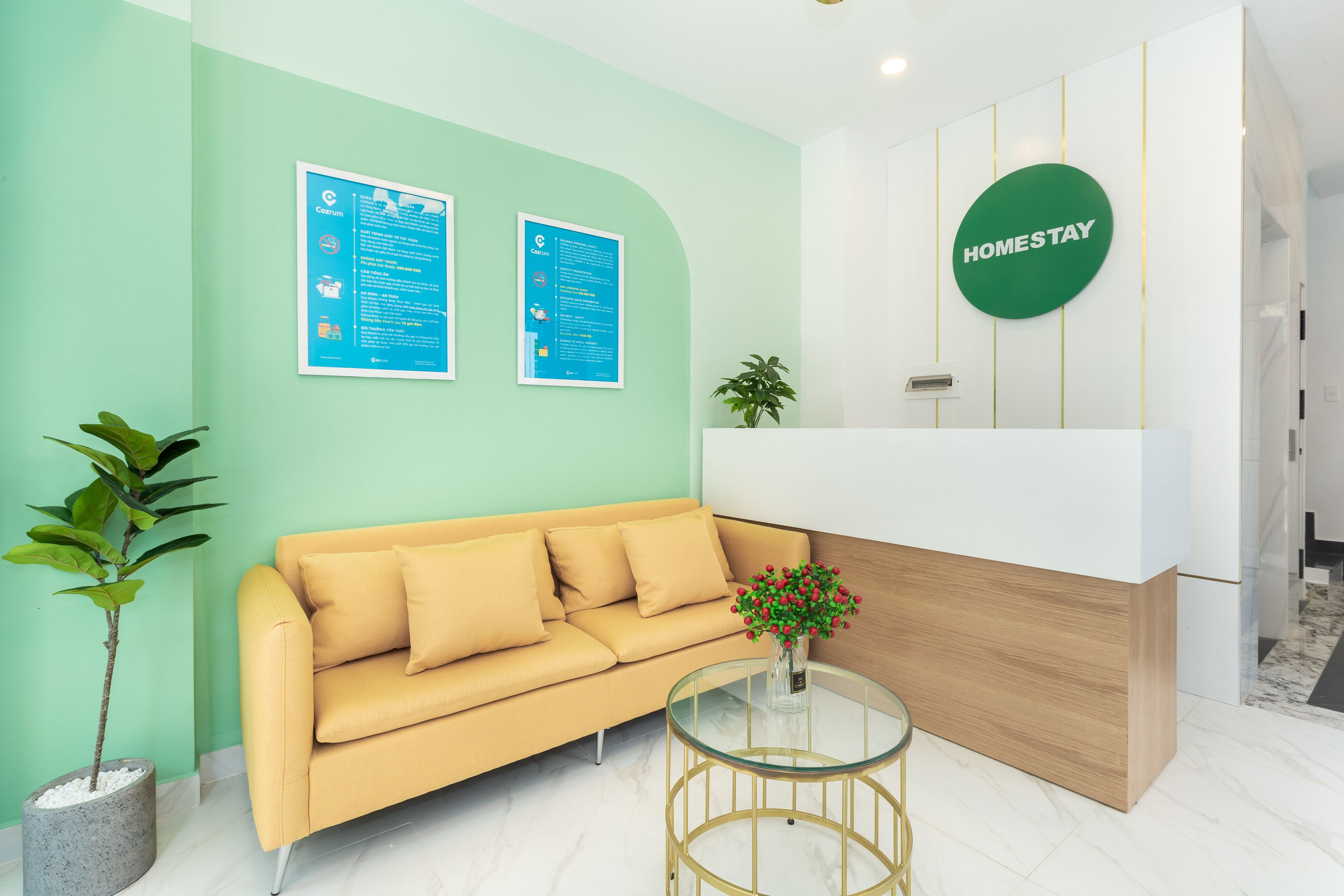 Un área de recepción con una pared verde, un mostrador de recepción, un sofá amarillo, una mesa de vidrio con un jarrón de flores y una planta en maceta.