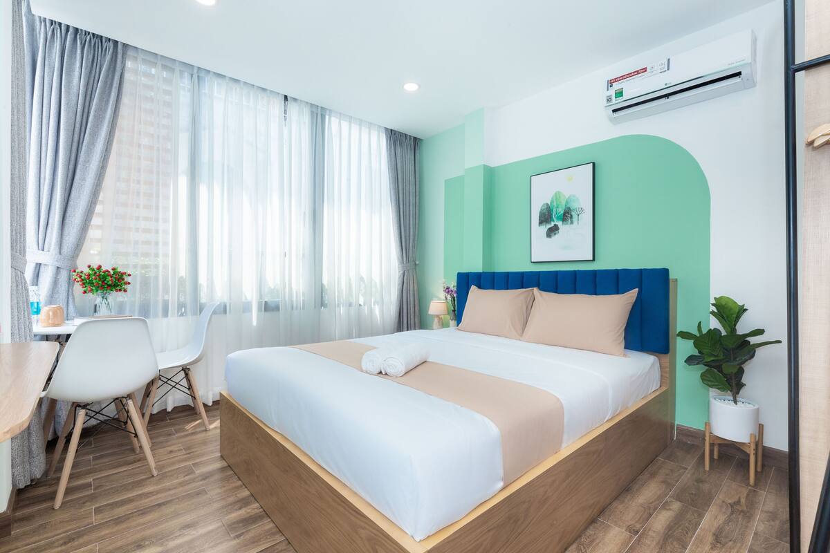 Een moderne hotelkamer met een groot bed, een bureau met een stoel, een kleine tafel met een bloevas en een kamerplant.
