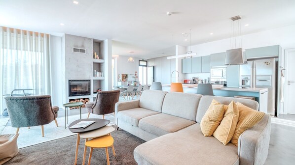 A modern living room with a sofa, chairs, a fireplace, and a kitchen in the background.