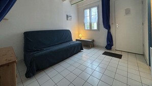 Pieni, niukasti kalustettu huone, jossa on yksi sänky, penkki, ikkuna, jossa on verhot, ja ovi.