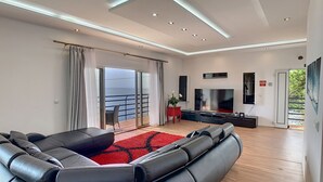 A modern living room with a leather sofa, a flat-screen TV, and a balcony with a view of the sea. - Villa Prainha by LovelyStay (Canical)