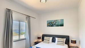 A bedroom with a bed, two bedside tables, a window with curtains, a painting on the wall, and a view of the outdoors. - Villa Prainha by LovelyStay (Canical)