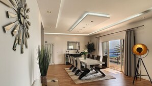 A modern dining room with a glass table, wooden flooring, and a view of the sea through large windows. - Villa Prainha by LovelyStay (Canical)