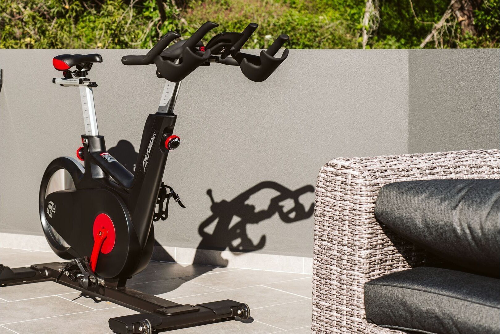 Una cyclette da esterno con sella rossa, una poltrona in vimini e un muro proiettano un'ombra.