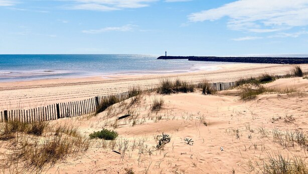 Une plage bordée d’une clôture en bois, avec des dunes de sable et un phare visible au loin.