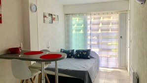 A small, well-lit room with a bed, a table with two red coasters, a white chair, and a window with blinds. - Cozy Air-Conditioned Studio with Terrace & Parking, Ideal for 2 in Vibrant Agde (Cap d'Agde naturisme)
