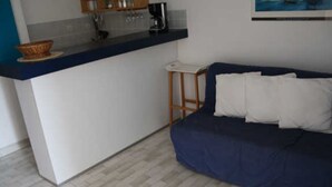 A compact living space with a kitchenette, a sofa with pillows, and a small wooden stool. - Apartment for 4, equipped kitchen, terrace, parking, Cap d'Agde (Cap d'Agde naturisme)
