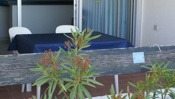 A room with a blue table and chairs, a wooden bench with a sign, and a view of a kitchen area. - Apartment for 4, equipped kitchen, terrace, parking, Cap d'Agde (Cap d'Agde naturisme)