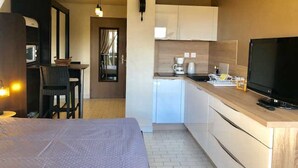 A hotel room with a bed, a desk with a chair, a kitchenette with a TV, and a bathroom with a shower. - Charming Studio with Pool Access, Terrace, and Parking in Agde (Cap d'Agde naturisme)