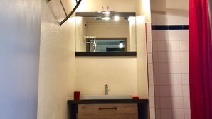 A bathroom with a wooden vanity, a mirror above the sink, and a red curtain. - Charming Studio with Pool Access, Terrace, and Parking in Agde (Cap d'Agde naturisme)