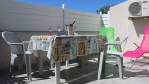 An outdoor patio with a table covered in a patterned tablecloth, surrounded by plastic chairs, one of which is pink. - Cozy Air-Conditioned Apartment with Pool Access, Terrace & Private Parking in Agde (Cap d'Agde naturisme)