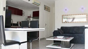 A living room with a black sofa, a glass coffee table, and a white desk with a computer. There is a kitchen in the background with red cabinets and a microwave. - Cozy Air-Conditioned Apartment with Pool Access, Terrace & Private Parking in Agde (Cap d'Agde naturisme)