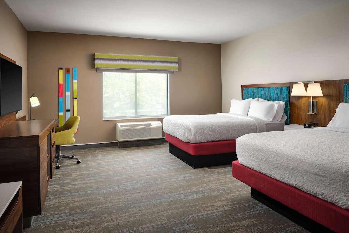A hotel room with two beds, a desk, a chair, a TV, a window, and a wall with colorful decor.