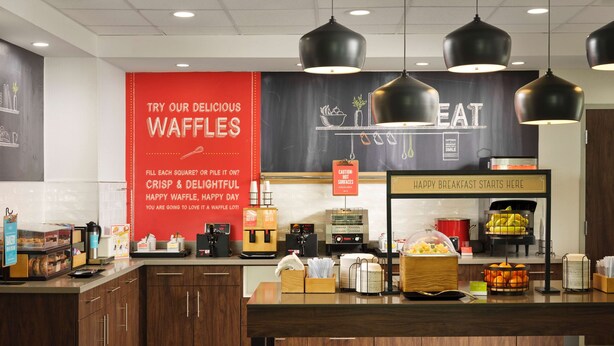 Un espace dédié au petit-déjeuner, comprenant un coin où l’on prépare des gaufres, des machines à café et un panneau publicitaire faisant la promotion des gaufres.