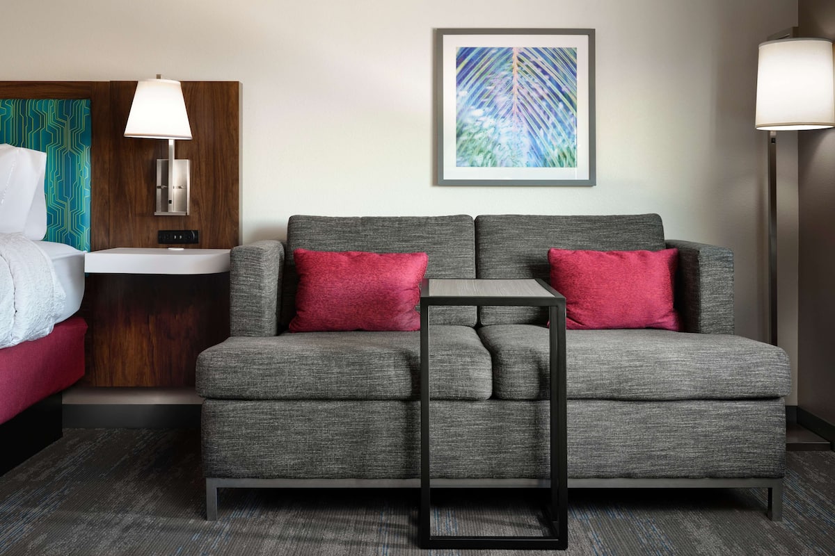 A modern hotel room with a grey sofa, red pillows, a small table, a bedside table with a lamp, a framed artwork on the wall, and a bathroom with a towel rack.