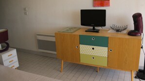 A room with a wooden desk, a black monitor, and a red hairdryer. There is a white radiator on the wall and a white cabinet with drawers.