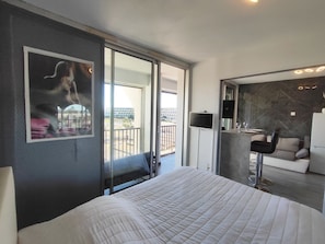 Habitación de hotel con una cama grande, televisor, balcón con vistas y un pequeño rincón de descanso con sofá y mesa.