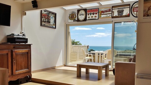 Une pièce baignée de lumière, avec un meuble en bois, une table et des chaises. Une grande porte vitrée donne accès à un espace extérieur offrant une vue sur la mer.