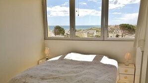 Une chambre à coucher avec un lit, deux tables de chevet avec des lampes, une fenêtre donnant sur la mer et les bâtiments, et une porte.
